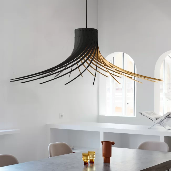 Grande suspension design en bois noir et intérieur doré en forme de jupe évasée ou de chapeau suspendu au-dessus d'une table de salle à manger. Les lamelles de bois s'écartent avec élégance pour laisser passer la lumière. En arrière-plan, une pièce épurée blanche avec de grandes fenêtres voûtées, un buffet intégré, deux verres ambrés et un pichet en terre cuite sur la table.